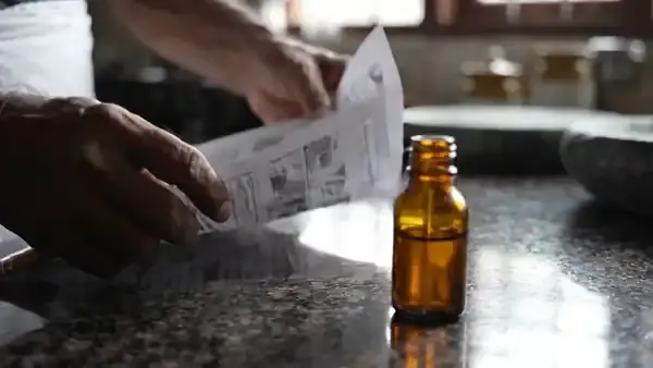 A man's hands smoothing out a printed sheet on a stone counter and running a pencil down the margin beside a small amber bottle
